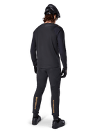 Achteraanzicht van een mountainbiker die het zwarte Alpinestars A-Aria Polartec Switch Jersey met lange mouwen draagt, gecombineerd met een zwarte MTB-broek met lichtbruine Alpinestars-branding op de kuiten, zwarte helm en handschoenen, professionele wieleruitrusting voor off-road paden