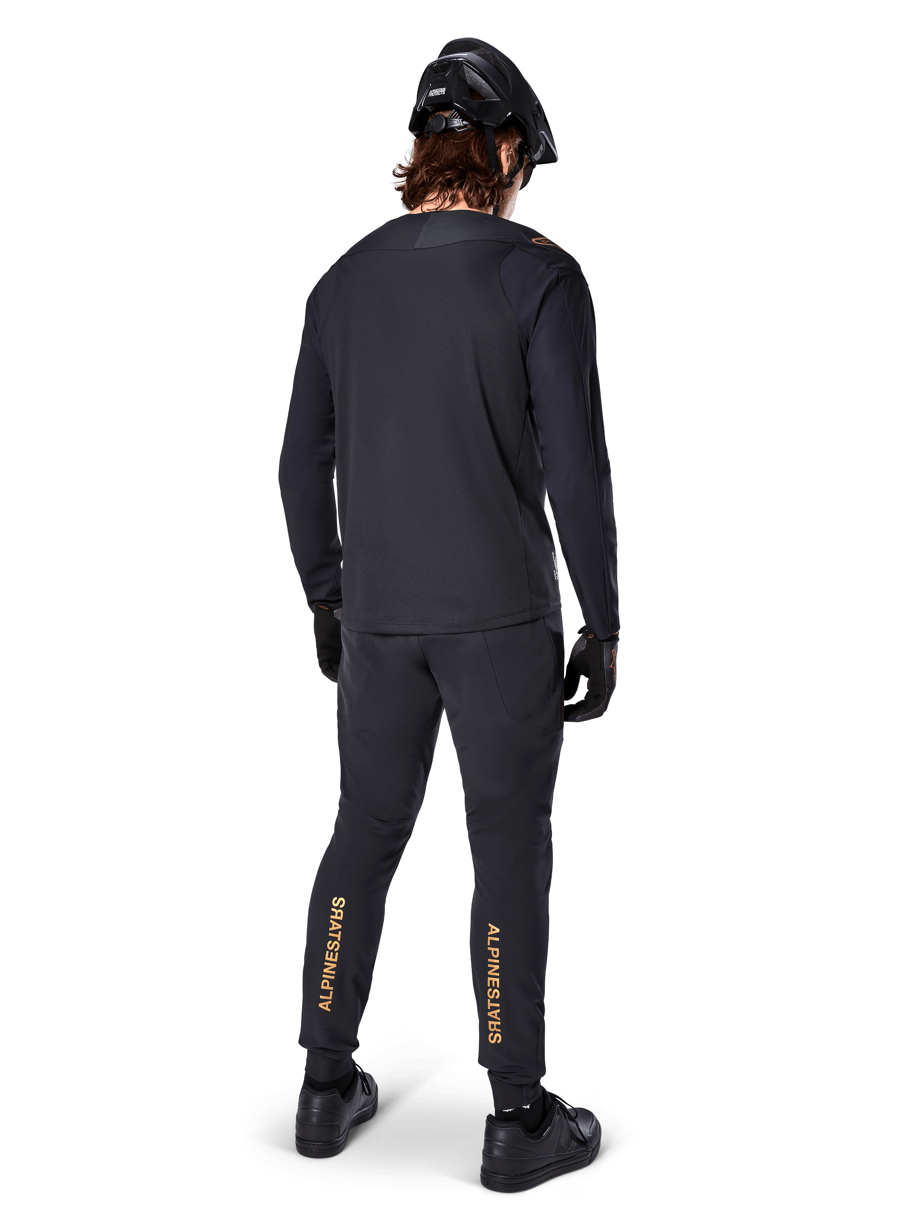 Achteraanzicht van een mountainbiker die het zwarte Alpinestars A-Aria Polartec Switch Jersey met lange mouwen draagt, gecombineerd met een zwarte MTB-broek met lichtbruine Alpinestars-branding op de kuiten, zwarte helm en handschoenen, professionele wieleruitrusting voor off-road paden