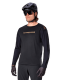 Alpinestars A-Aria Polartec Switch fietsshirt met lange mouwen, zwart met goudkleurig logo, MTB mountainbike-kleding, gedragen met een zwarte helm en zonnebril, met lasergesneden ventilatie op de mouwen en vochtafvoerende Polartec-stof voor off-road prestaties