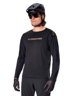 Alpinestars A-Aria Polartec Switch fietsshirt met lange mouwen, zwart met goudkleurig logo, MTB mountainbike-kleding, gedragen met een zwarte helm en zonnebril, met lasergesneden ventilatie op de mouwen en vochtafvoerende Polartec-stof voor off-road prestaties