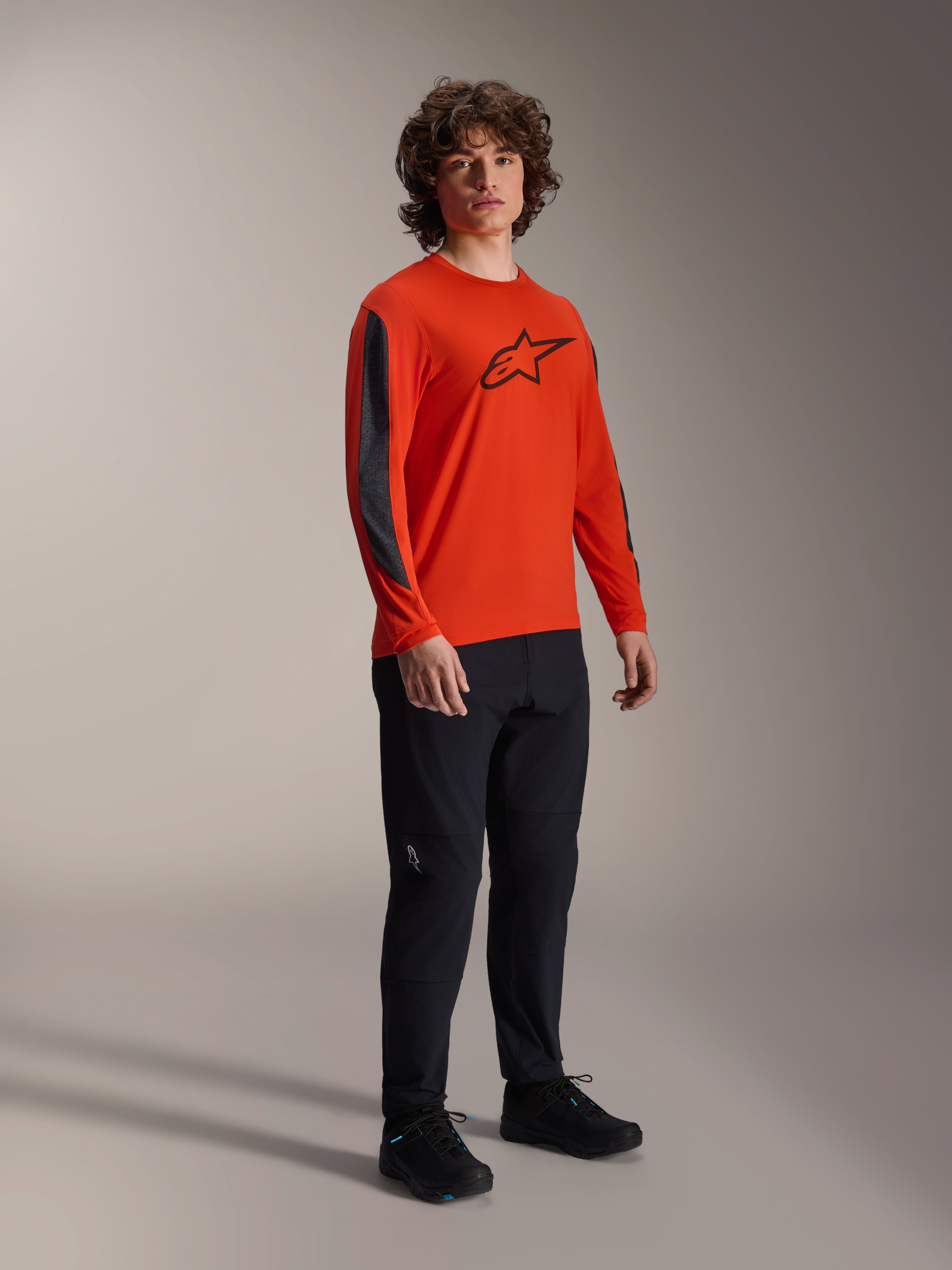 Mannelijk model draagt Alpinestars A-Dura Insert Astar Jersey met lange mouwen, Spicy Orange met zwart Astar-logo op de borst, donkergrijze mouwinzetten, zwarte broek, professionele MTB-wielerkleding