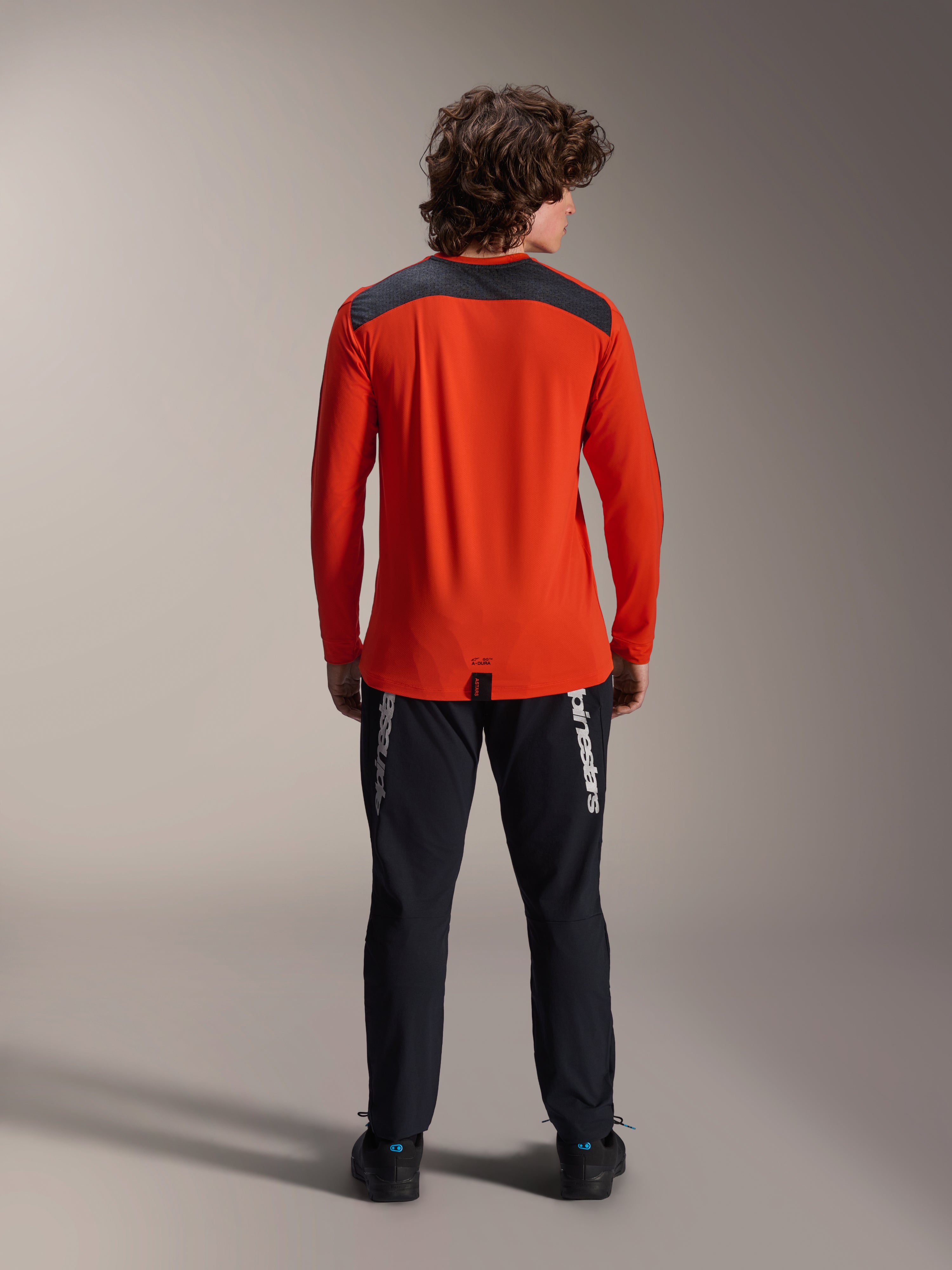 Alpinestars A-Dura Insert Astar Jersey met lange mouwen in Spicy Orange, achteraanzicht met antracietgrijs schouderpaneel, gedragen met zwarte Alpinestars-broek en zwarte schoenen, professionele studiofoto van MTB-prestatiekleding