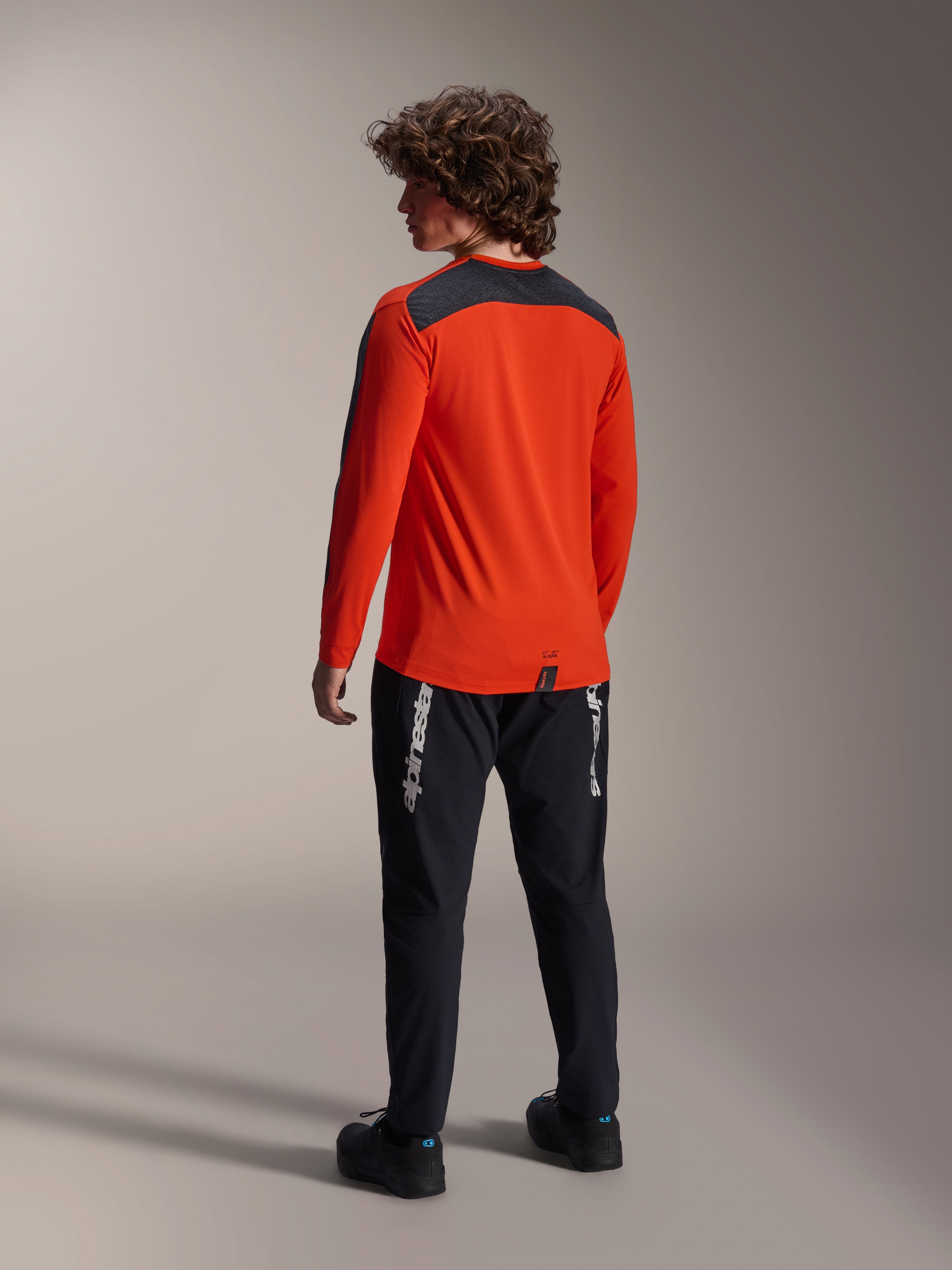 Achteraanzicht van een model dat de Alpinestars A-Dura Insert Astar Jersey in Spicy Orange draagt, MTB-jersey met lange mouwen en een donkergrijs schouderpaneel, gecombineerd met een zwarte merkbroek, off-road fietskleding.
