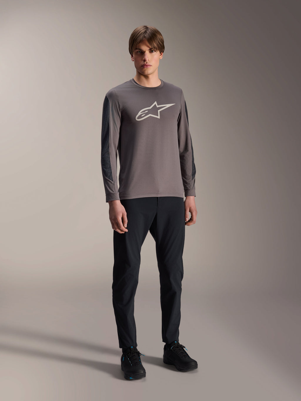 Alpinestars A-Dura Insert Astar Jersey, MTB-shirt met lange mouwen, donkergrijs, met groot wit logo op de borst, gedragen met zwarte technische broek en zwarte schoenen, off-road fietskleding