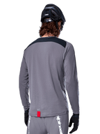 Alpinestars A-Dura Dri Astar Long Sleeve Jersey, Dark Gray MTB jersey shown from the back, featuring a black shoulder panel and small red brand tag, worn with a black mountain bike helmet and gloves.