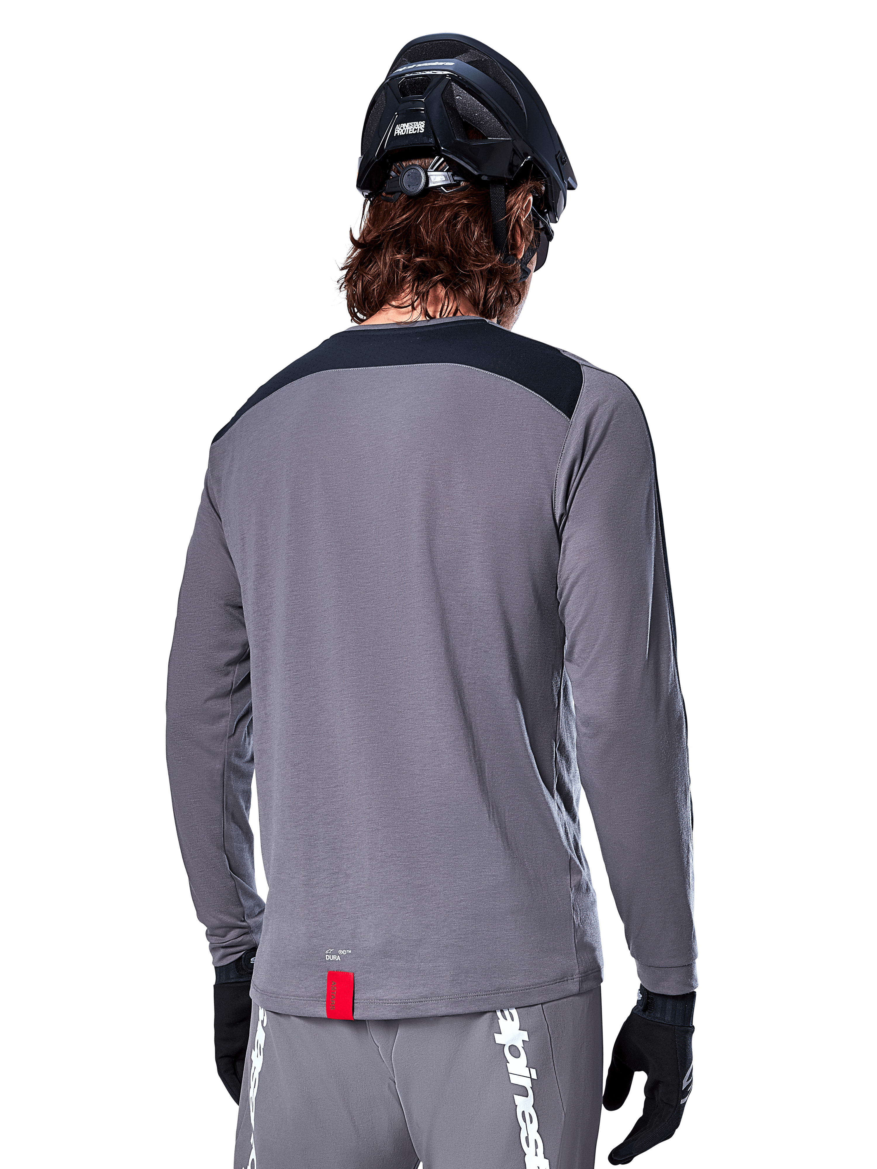 Alpinestars A-Dura Dri Astar Long Sleeve Jersey, Dark Gray MTB jersey shown from the back, featuring a black shoulder panel and small red brand tag, worn with a black mountain bike helmet and gloves.