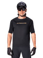 Alpinestars A-Aria Polartec Switch jersey met korte mouwen, zwart met goudkleurige branding op de borst, mountainbiker met zwarte MTB-helm, donkere sportzonnebril en bijpassende handschoenen, off-road fietskleding