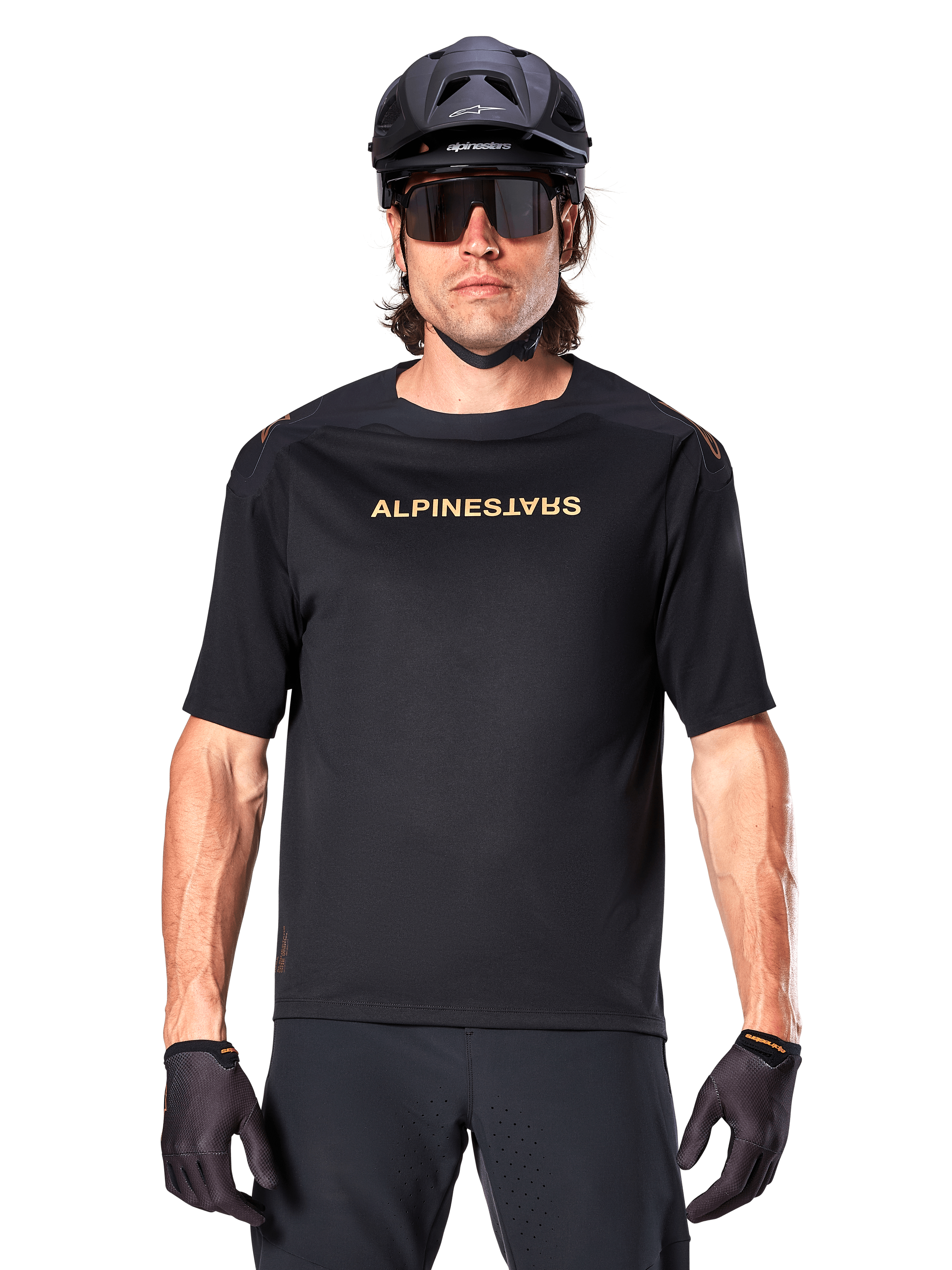 Alpinestars A-Aria Polartec Switch jersey met korte mouwen, zwart met goudkleurige branding op de borst, mountainbiker met zwarte MTB-helm, donkere sportzonnebril en bijpassende handschoenen, off-road fietskleding
