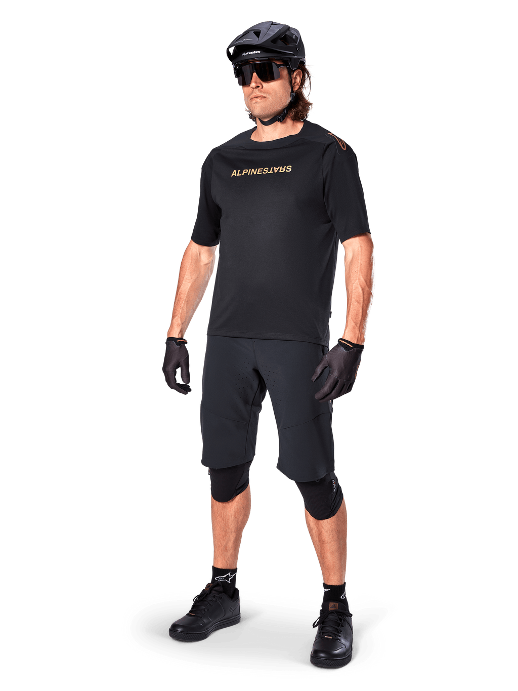 Alpinestars A-Aria Polartec Switch jersey met korte mouwen, zwart, met gespiegeld gouden logo op de borst, gedragen door een rijder met zwarte fietsshorts, helm en kniebeschermers, technische MTB-uitrusting voor off-road prestaties