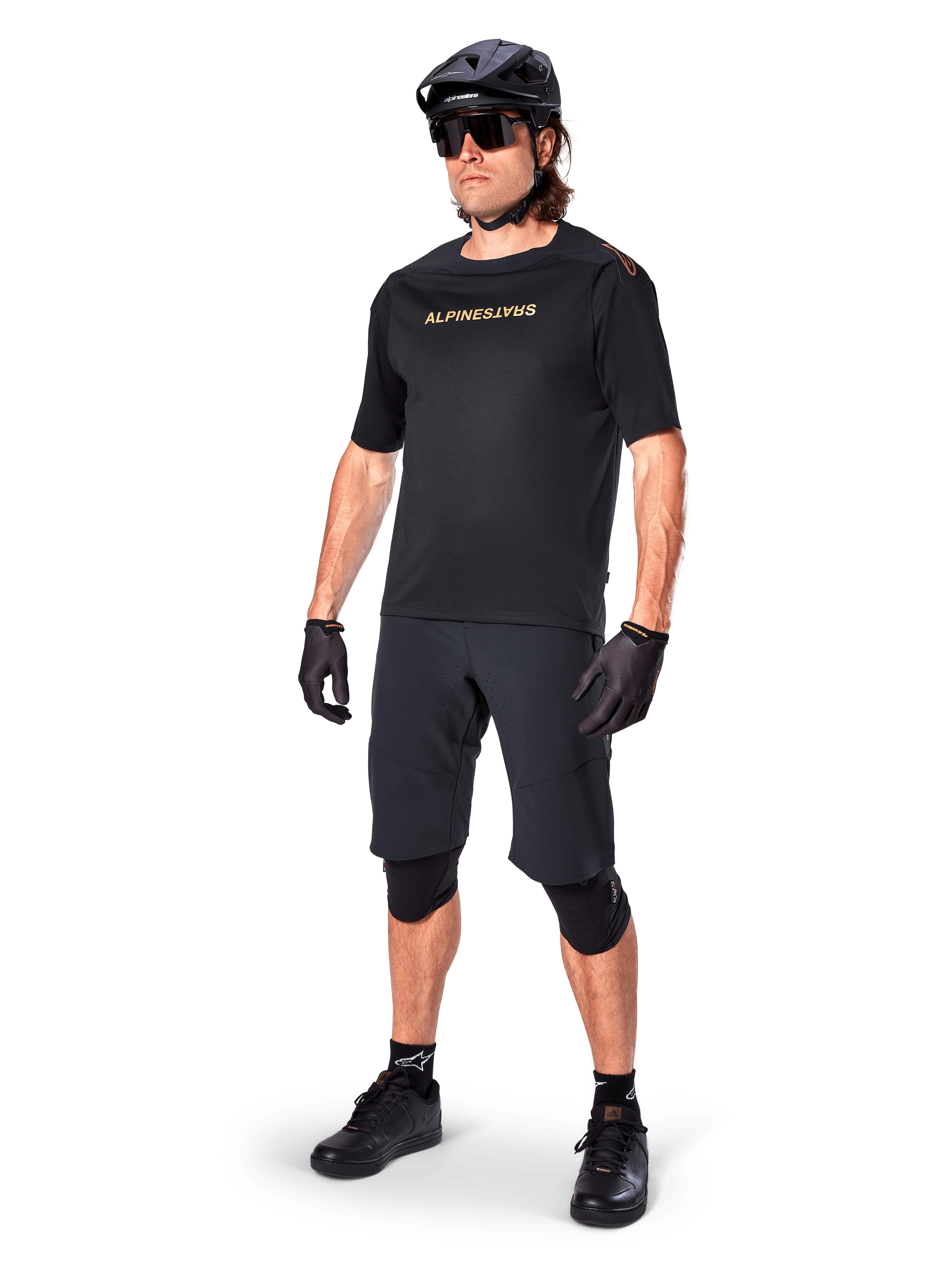 Alpinestars A-Aria Polartec Switch jersey met korte mouwen, zwart, met gespiegeld gouden logo op de borst, gedragen door een rijder met zwarte fietsshorts, helm en kniebeschermers, technische MTB-uitrusting voor off-road prestaties
