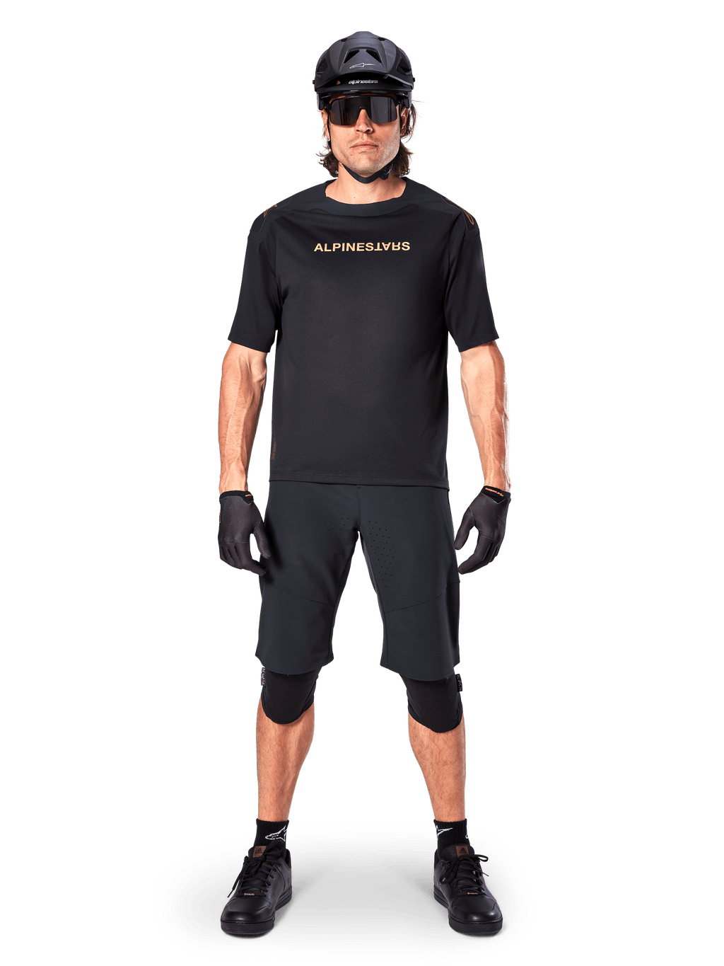 Alpinestars A-Aria Polartec Switch Jersey, MTB-shirt met korte mouwen, zwart met lichtbruine letters, gedragen met bijpassende zwarte mountainbike-uitrusting inclusief helm, short en kniebeschermers, hoogwaardige off-road fietskleding