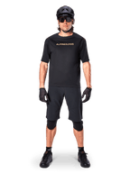 Alpinestars A-Aria Polartec Switch Jersey, MTB-shirt met korte mouwen, zwart met lichtbruine letters, gedragen met bijpassende zwarte mountainbike-uitrusting inclusief helm, short en kniebeschermers, hoogwaardige off-road fietskleding