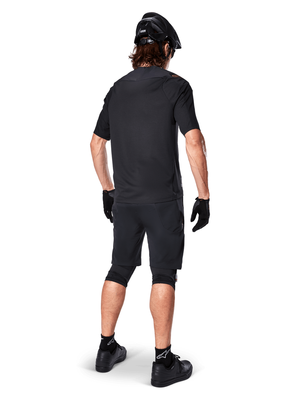 Mannelijke mountainbiker van achteren gezien die de Alpinestars A-Aria Polartec Switch Jersey in het zwart draagt, MTB-shirt met korte mouwen, gecombineerd met een zwarte mountainbike short, zwarte helm en handschoenen, gecoördineerde performance-outfit voor off-road trailrijden