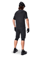 Mannelijke mountainbiker van achteren gezien die de Alpinestars A-Aria Polartec Switch Jersey in het zwart draagt, MTB-shirt met korte mouwen, gecombineerd met een zwarte mountainbike short, zwarte helm en handschoenen, gecoördineerde performance-outfit voor off-road trailrijden