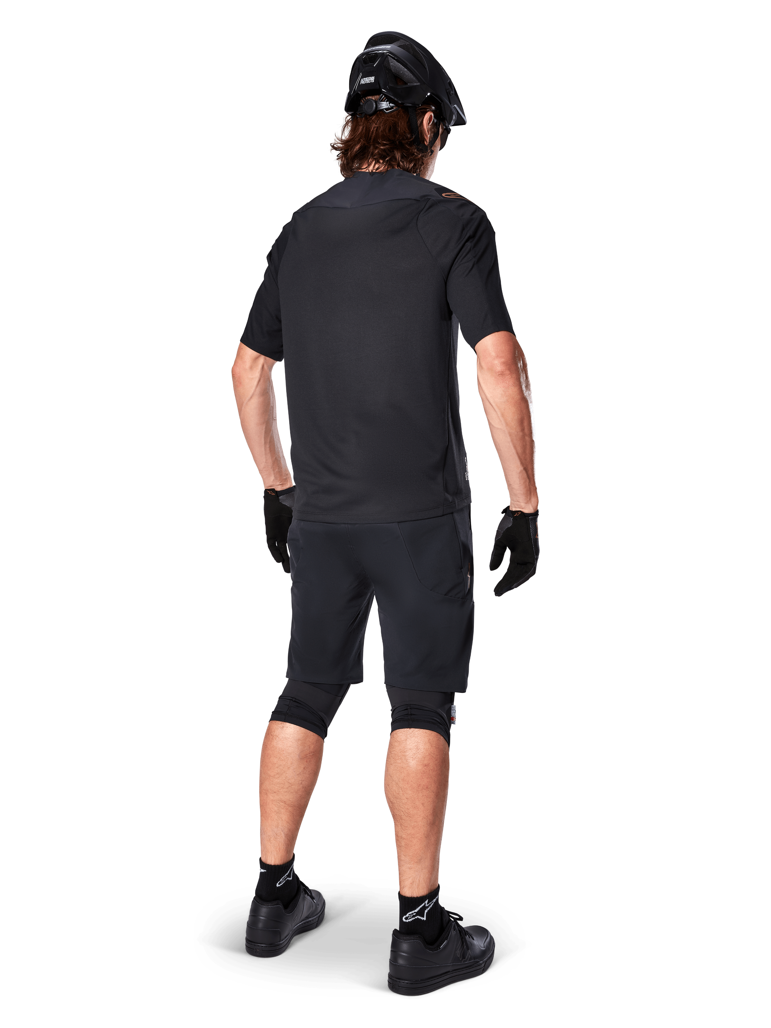 Mannelijke mountainbiker van achteren gezien die de Alpinestars A-Aria Polartec Switch Jersey in het zwart draagt, MTB-shirt met korte mouwen, gecombineerd met een zwarte mountainbike short, zwarte helm en handschoenen, gecoördineerde performance-outfit voor off-road trailrijden