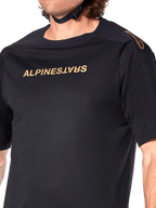 Alpinestars A-Aria Polartec Switch Jersey, MTB-jersey met korte mouwen, zwart, gouden Alpinestars-belettering op de borst en logo op de schouder, close-up van het bovenlichaam met focus op de technische stof en prestatiegerichte pasvorm