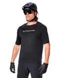 Alpinestars A-Aria Polartec Switch jersey met korte mouwen, zwart mountainbike-shirt met goudkleurige Alpinestars-branding, gedragen met zwarte helm, zonnebril en handschoenen, technische MTB-kleding