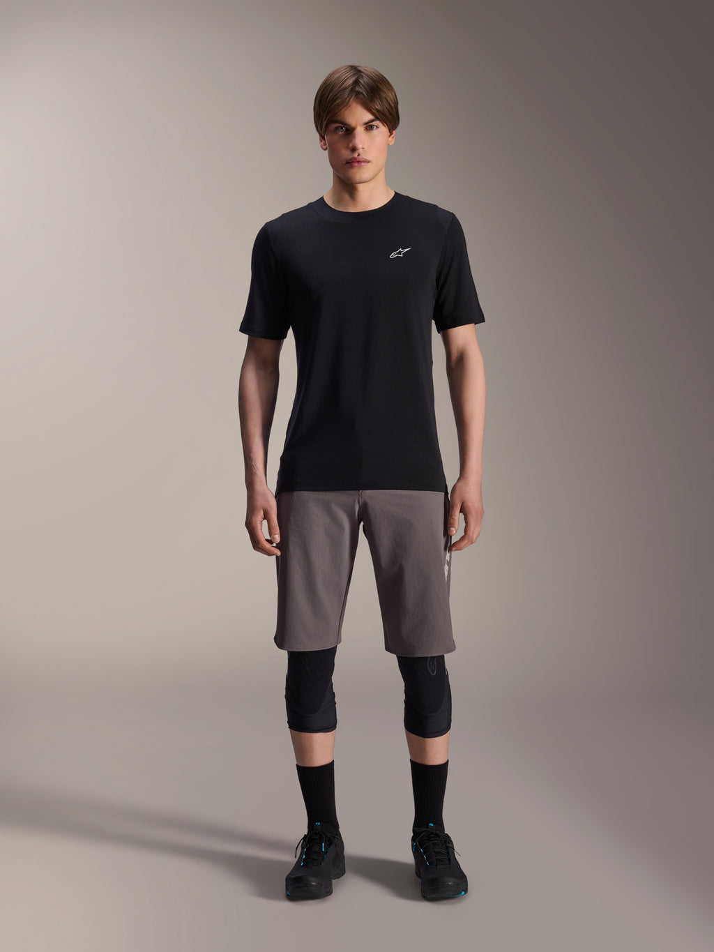 Mannelijk model draagt Alpinestars A-Dura Elite Shorts in donkergrijs en A-Dura Dri-Wool Jersey met korte mouwen in zwart, gecombineerd met zwarte kniebeschermers en fietsschoenen, technische mountainbikekleding voor off-road prestaties.