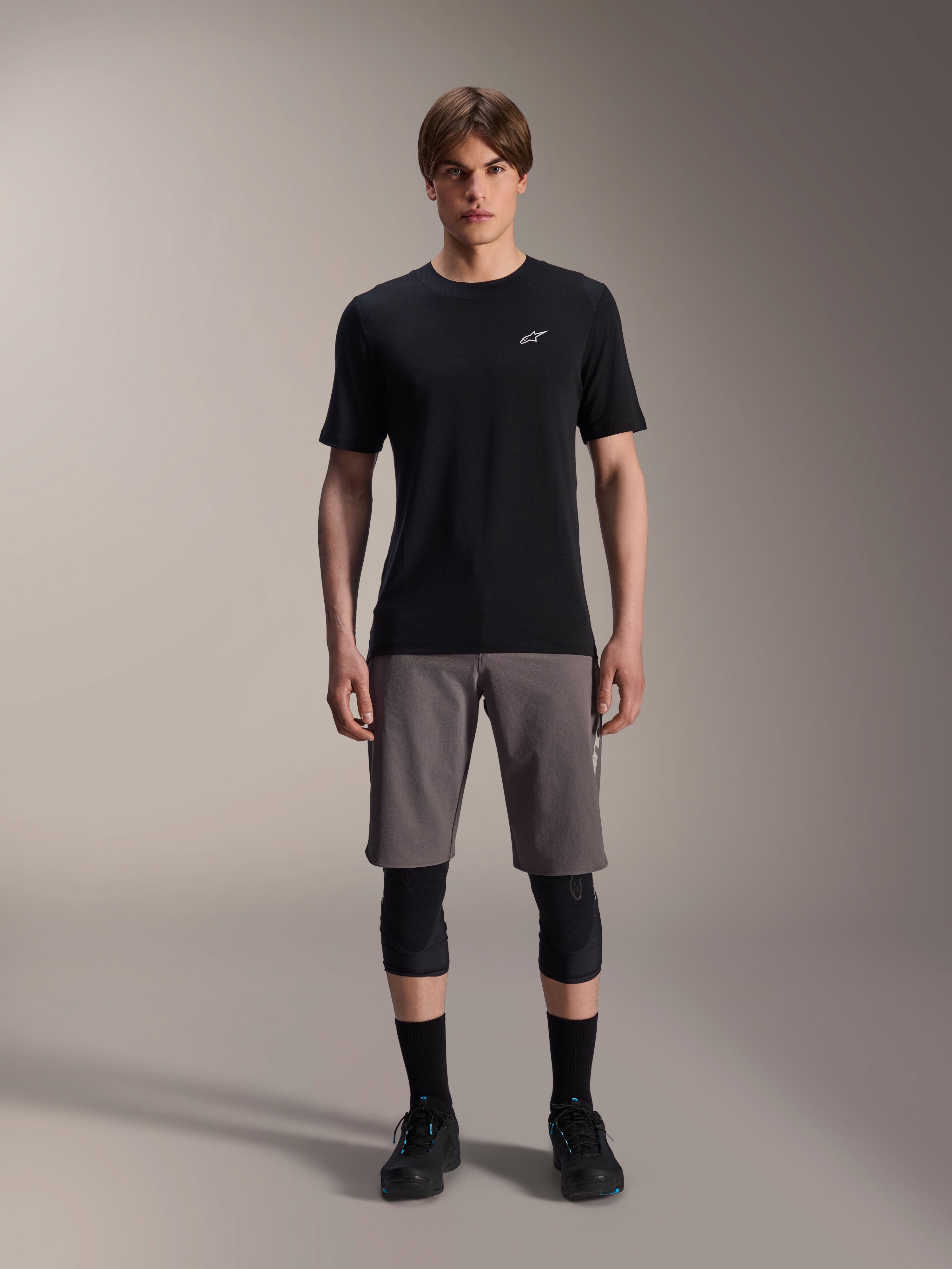 Alpinestars A-Dura Dri Wool Jersey met korte mouwen, zwart, gedragen door een model met grijze mountainbike shorts, zwarte kniebeschermers en zwarte fietsschoenen, minimalistisch ontwerp met een klein wit Astars-logo op de borst, functionele MTB-kleding voor trailrijden