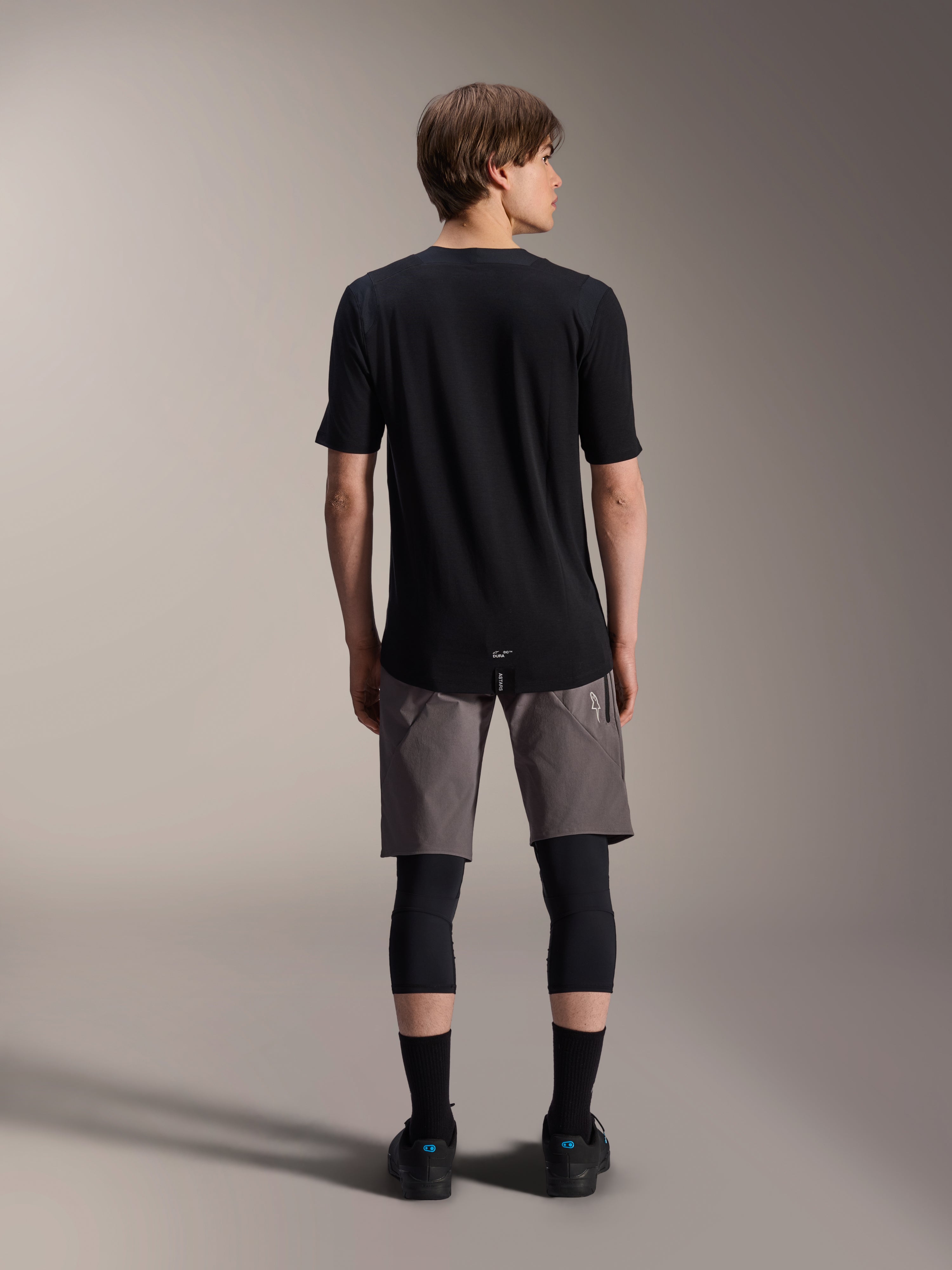 Alpinestars A-Dura Elite Shorts, donkergrijze mountainbike short, achteraanzicht, gedragen met een zwart jersey met korte mouwen, kniebeschermers en zwarte sokken, technische wielerkleding voor off-road rijden