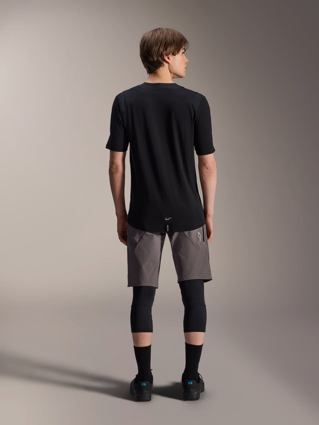 Alpinestars A-Dura Dri Wool Jersey met korte mouwen, zwart, achteraanzicht gedragen door model met grijze MTB-short en zwarte kniebeschermers, voorzien van een langere achterzijde en atletische pasvorm voor off-road fietsen