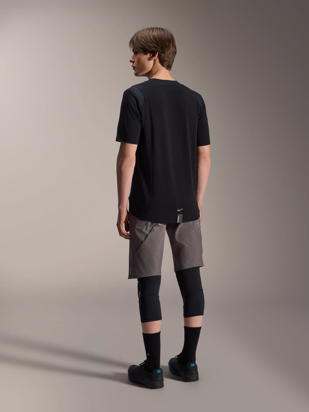 Alpinestars A-Dura Dri Wool Jersey met korte mouwen en A-Dura Elite Shorts, zwarte en donkergrijze mountainbikekleding, gedragen met zwarte kniebeschermers en sokken, achteraanzicht van fietskleding voor off-road prestaties