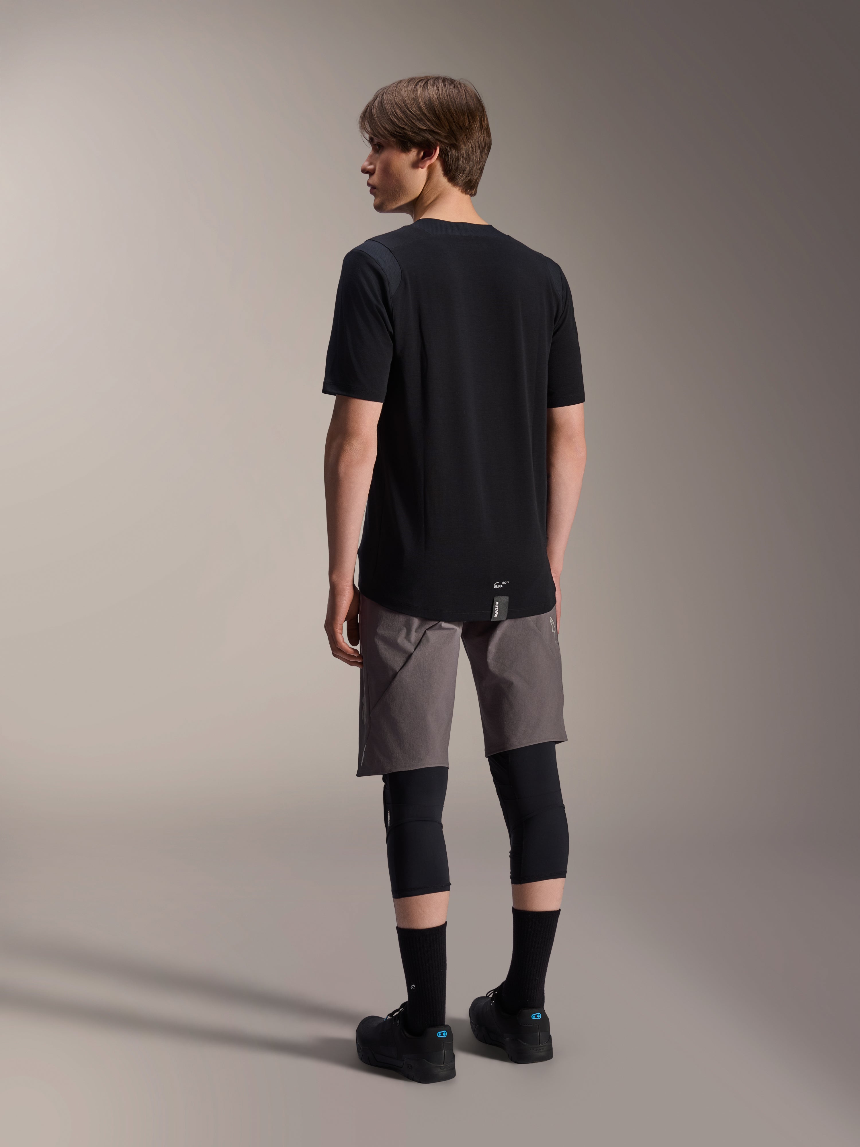 Alpinestars A-Dura Dri Wool Jersey met korte mouwen, zwart, achteraanzicht op model, gedragen met grijze mountainbike shorts en zwarte kniebeschermers, voorzien van een verlengde achterzijde en subtiel logo, technische MTB-kleding