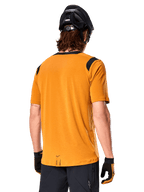 Alpinestars A-Dura Dri Wool Short Sleeve Jersey, Dark Gold, Rear view of a man wearing the short-sleeve jersey with black mesh panels on the shoulders, paired with a black MTB helmet and gloves, regular fit mountain bike gear.