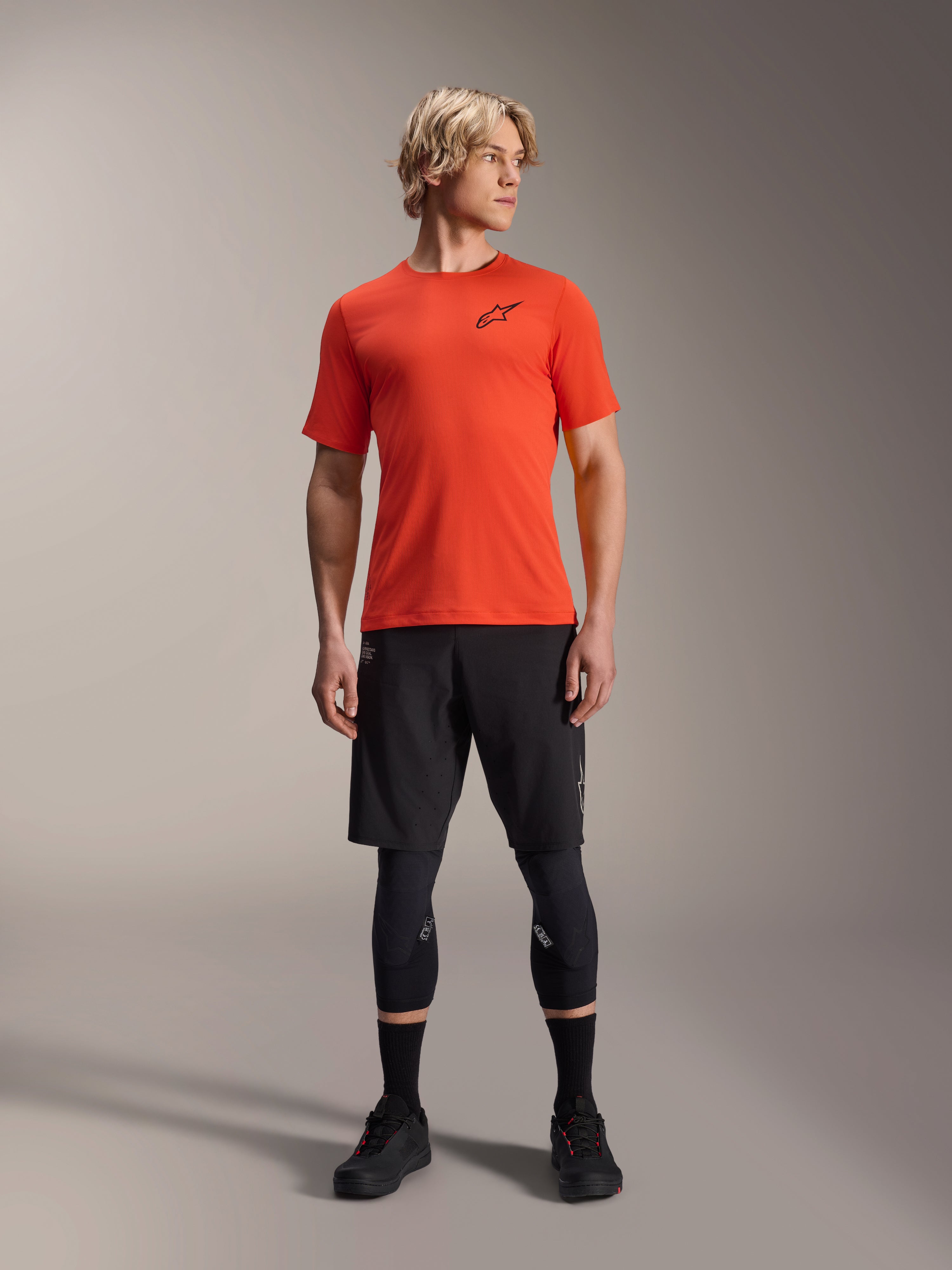 Alpinestars A-Aria Astar Jersey met korte mouwen, Spicy Orange, gedragen door model met zwarte mountainbike shorts en kniebeschermers, lichtgewicht en ademende performance fietskleding voor MTB