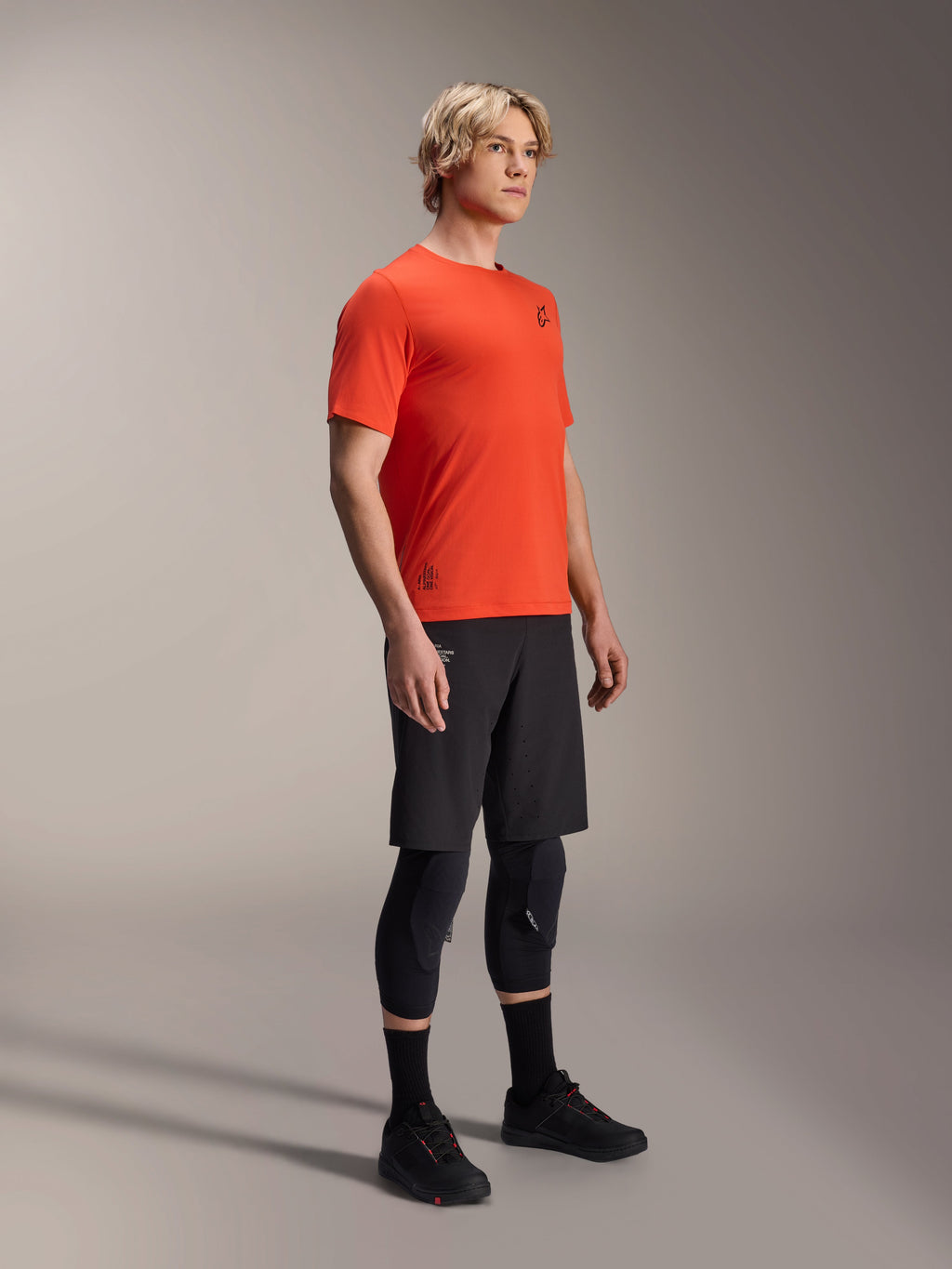 Alpinestars A-Aria Astar jersey met korte mouwen in Spicy Orange, gedragen door een model met een zwarte MTB-short en kniebescherming, lichtgewicht ademende mountainbikekleding, levendig oranje met minimale zwarte branding
