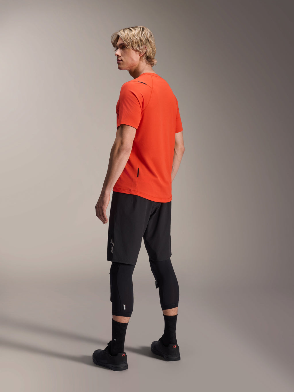 Alpinestars A-Aria Astar jersey met korte mouwen, Spicy Orange, driekwart achteraanzicht van model dat over de schouder kijkt, gecombineerd met zwarte MTB-shorts en kniebeschermers, technische mountainbikekleding