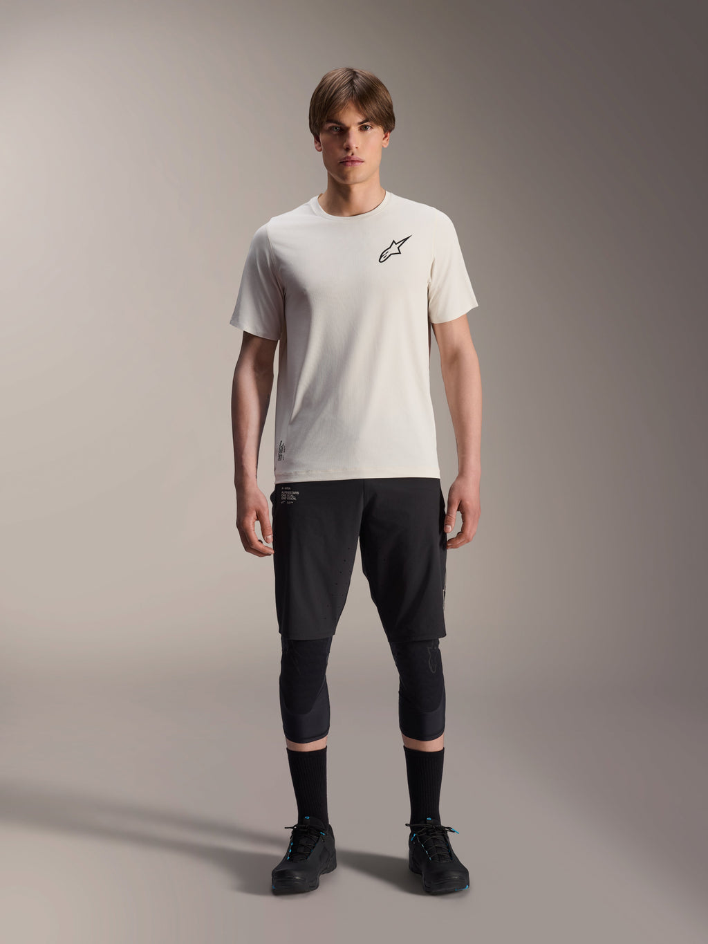 Alpinestars A-Aria Astar Jersey met korte mouwen in Ecru, mannelijk model draagt zwarte MTB-shorts, kniebeschermers en zwarte fietsschoenen, voorzien van een minimalistisch zwart sterlogo op de borst, ontworpen voor off-road trailrijden