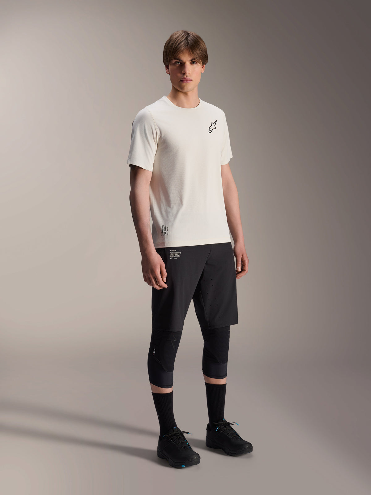 Alpinestars A-Aria Astar Jersey, korte mouwen, ecru, voorzien van een zwart logo op de borst, gedragen met een zwarte fietsbroek en kniebeschermers, lichtgewicht MTB-performancekleding
