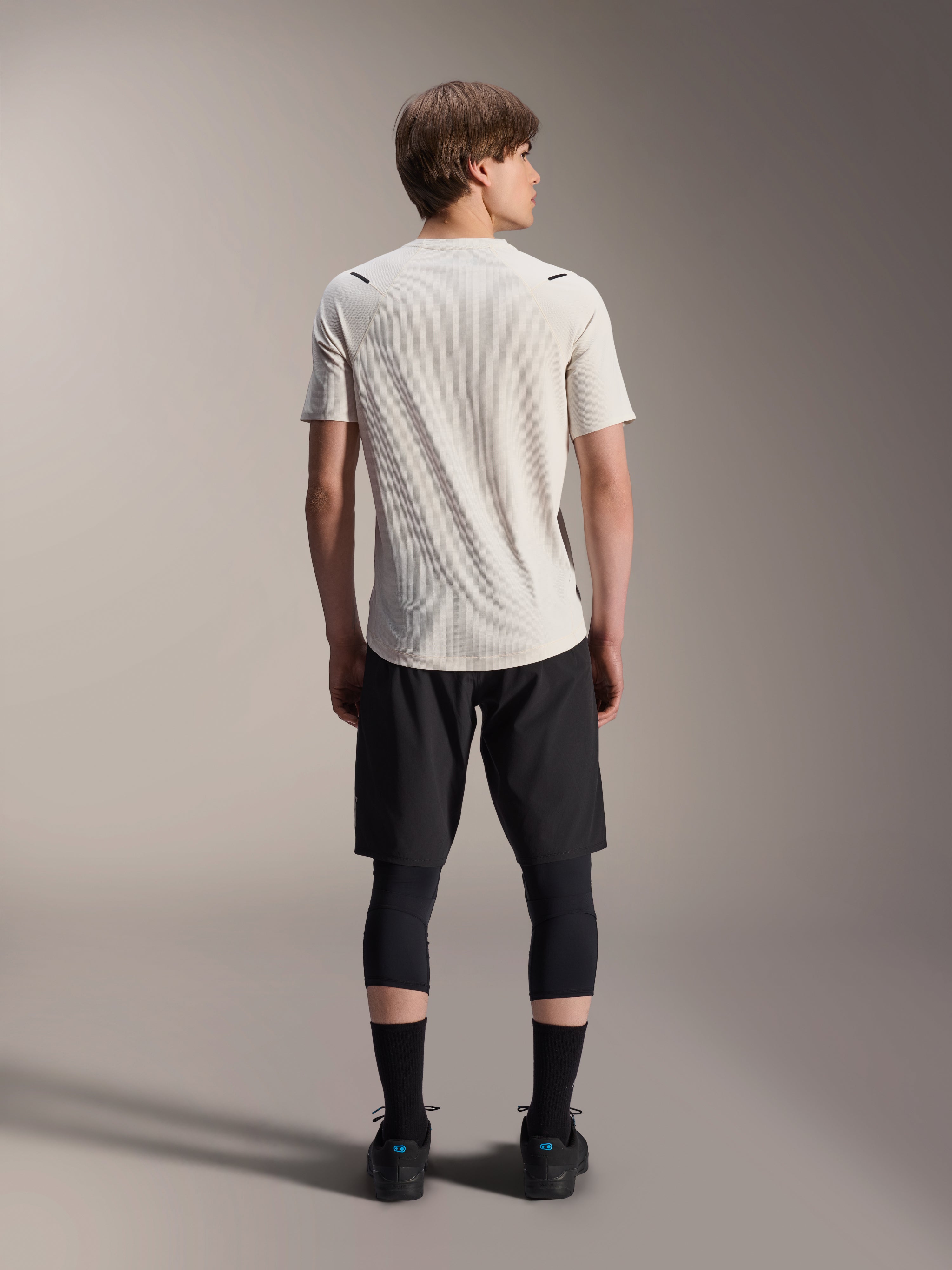 Mannelijk model vanaf de achterkant, gekleed in het Alpinestars A-Aria Astar jersey met korte mouwen in Ecru, gecombineerd met een zwarte MTB-short en beschermende leggings, voorzien van een verlengde achterkant en een prestatiepasvorm voor off-road trailrijden.