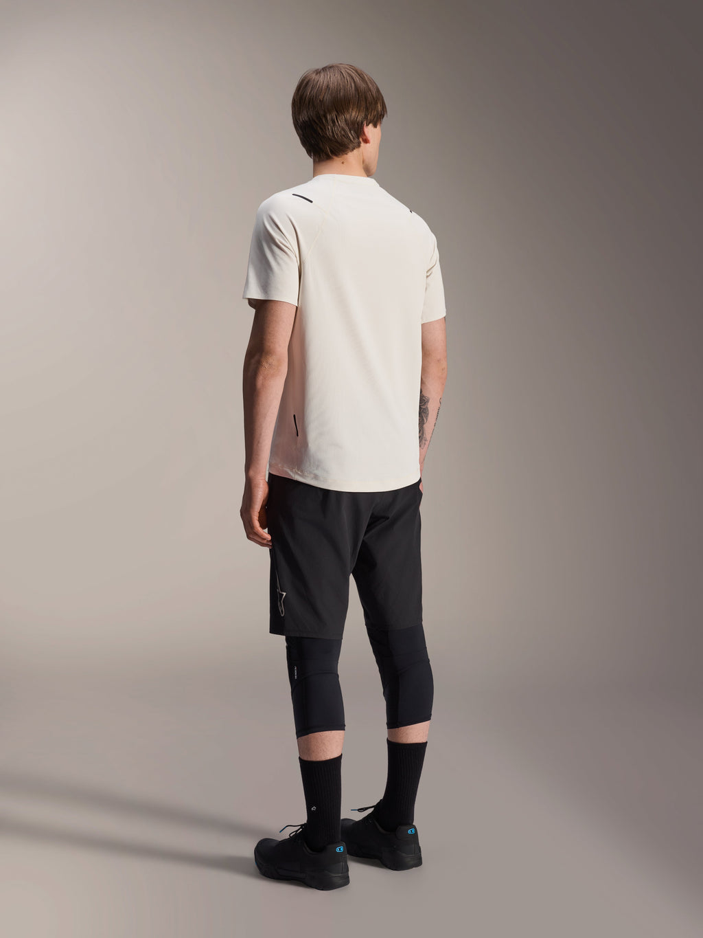 Alpinestars A-Aria Astar Jersey, MTB-shirt met korte mouwen, Ecru, achteraanzicht dat de prestatiegerichte pasvorm en verlengde achterzijde toont, gecombineerd met een zwarte mountainbike short, kniebeschermers en zwarte fietsschoenen voor off-road trailsessies