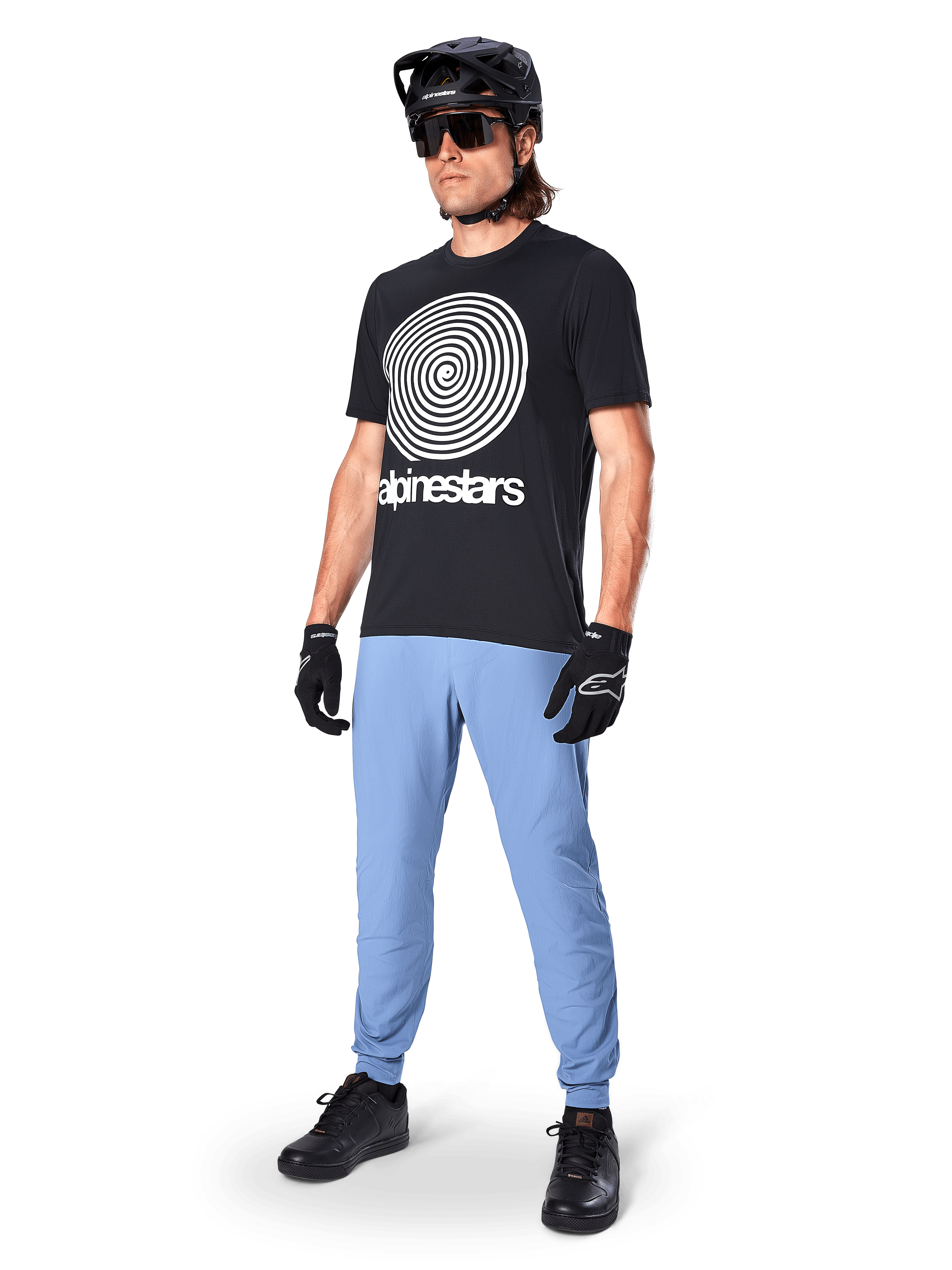 Alpinestars A-Dura Oscar Short Sleeve Jersey, Black MTB jersey featuring a large white heritage spiral graphic, worn by a rider with a black helmet, sunglasses, black gloves, and light blue trousers, performance mountain bike gear