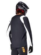 MTB-rijder draagt Alpinestars A-Supra Oscar jersey met lange mouwen, zwart met witte panelen op de schouders en mouwen, van achteren gezien, zwarte helm en handschoenen, broek met oranje accenten, duurzame off-road fietskleding