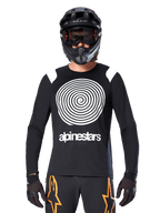 Alpinestars A-Supra Oscar Jersey met lange mouwen, zwart met witte spiraal-graphic en schouderaccenten, gedragen door een rijder met een zwarte integraalhelm en goggle, zwarte en oranje broek, hoogwaardige fietskleding voor MTB gravity rijden