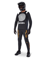 MTB-rijder draagt Alpinestars A-Supra Oscar Jersey met lange mouwen in het zwart, met de witte vintage Oscar-spiraalgrafiek, zwarte broek met oranje sterlogo's, zwarte full-face helm met goggle en zwarte handschoenen, professionele mountainbike-uitrusting