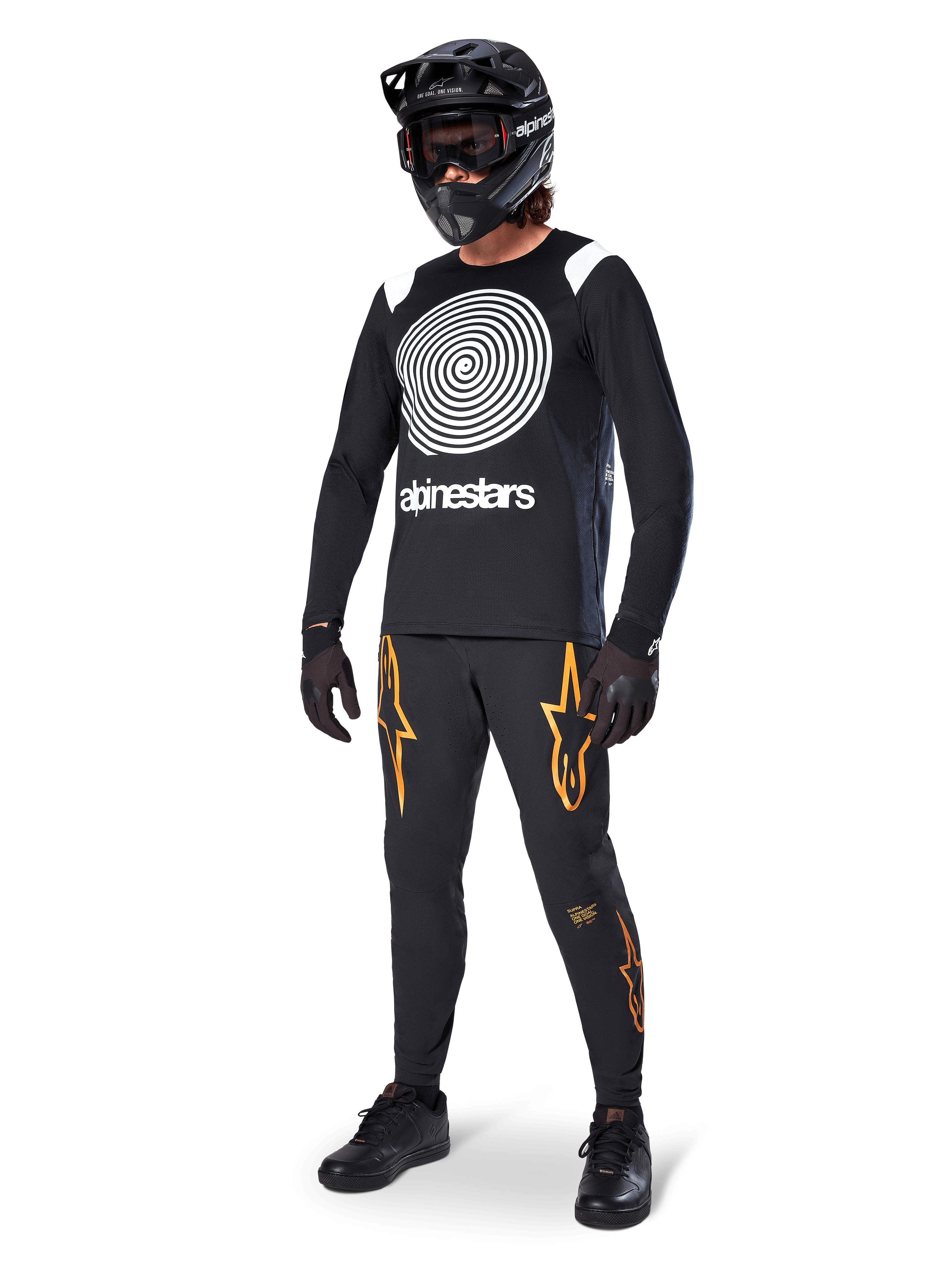MTB-rijder draagt Alpinestars A-Supra Oscar Jersey met lange mouwen in het zwart, met de witte vintage Oscar-spiraalgrafiek, zwarte broek met oranje sterlogo's, zwarte full-face helm met goggle en zwarte handschoenen, professionele mountainbike-uitrusting