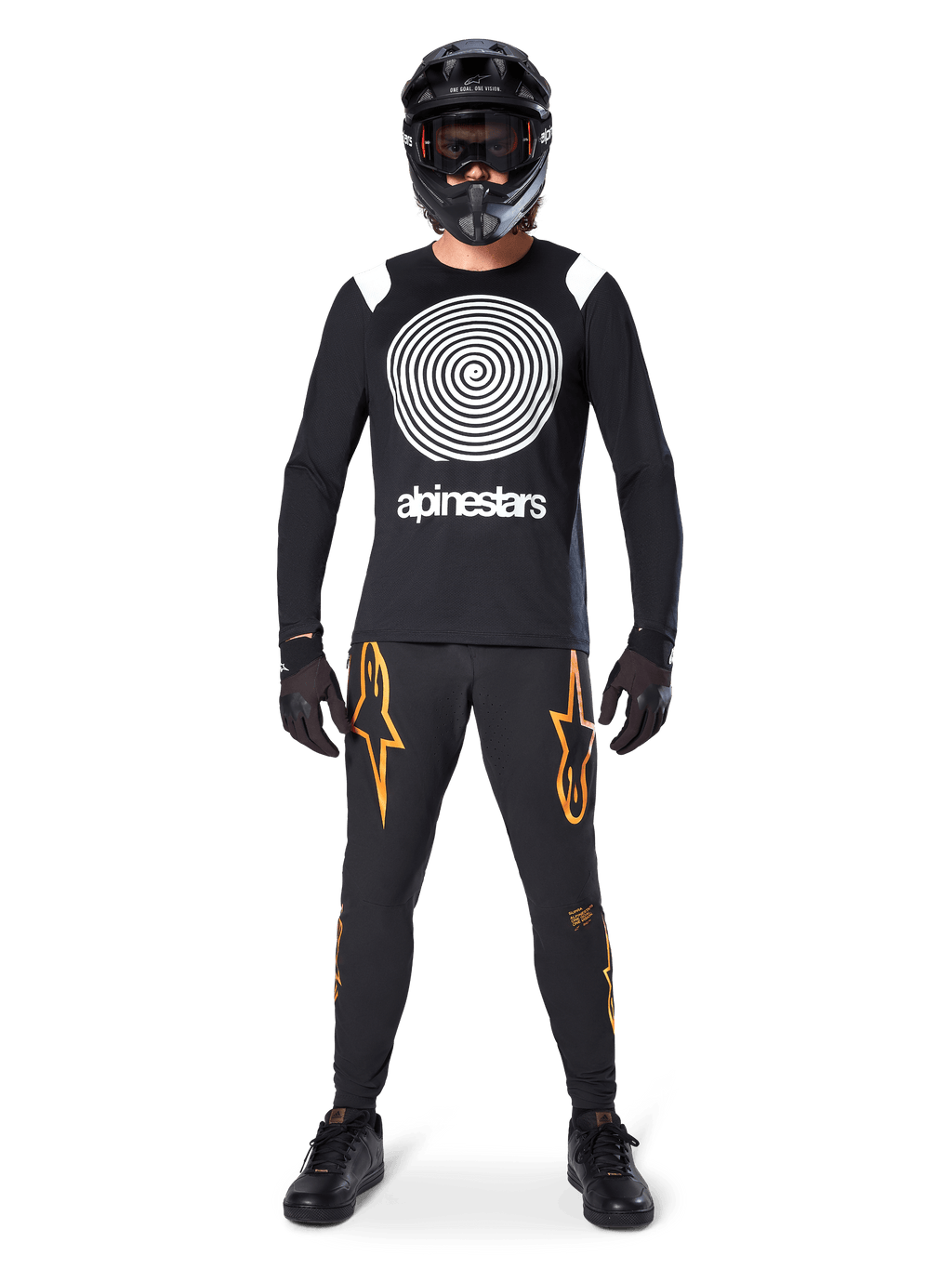 Mannelijk model in volledige mountainbike-uitrusting, Alpinestars A-Supra Oscar Jersey met lange mouwen in zwart met witte spiraalgrafiek op de borst, zwarte broek met oranje sterlogo's, zwarte full-face helm en bril, zwarte handschoenen en schoenen, gecoördineerde outfit voor MTB gravity riding