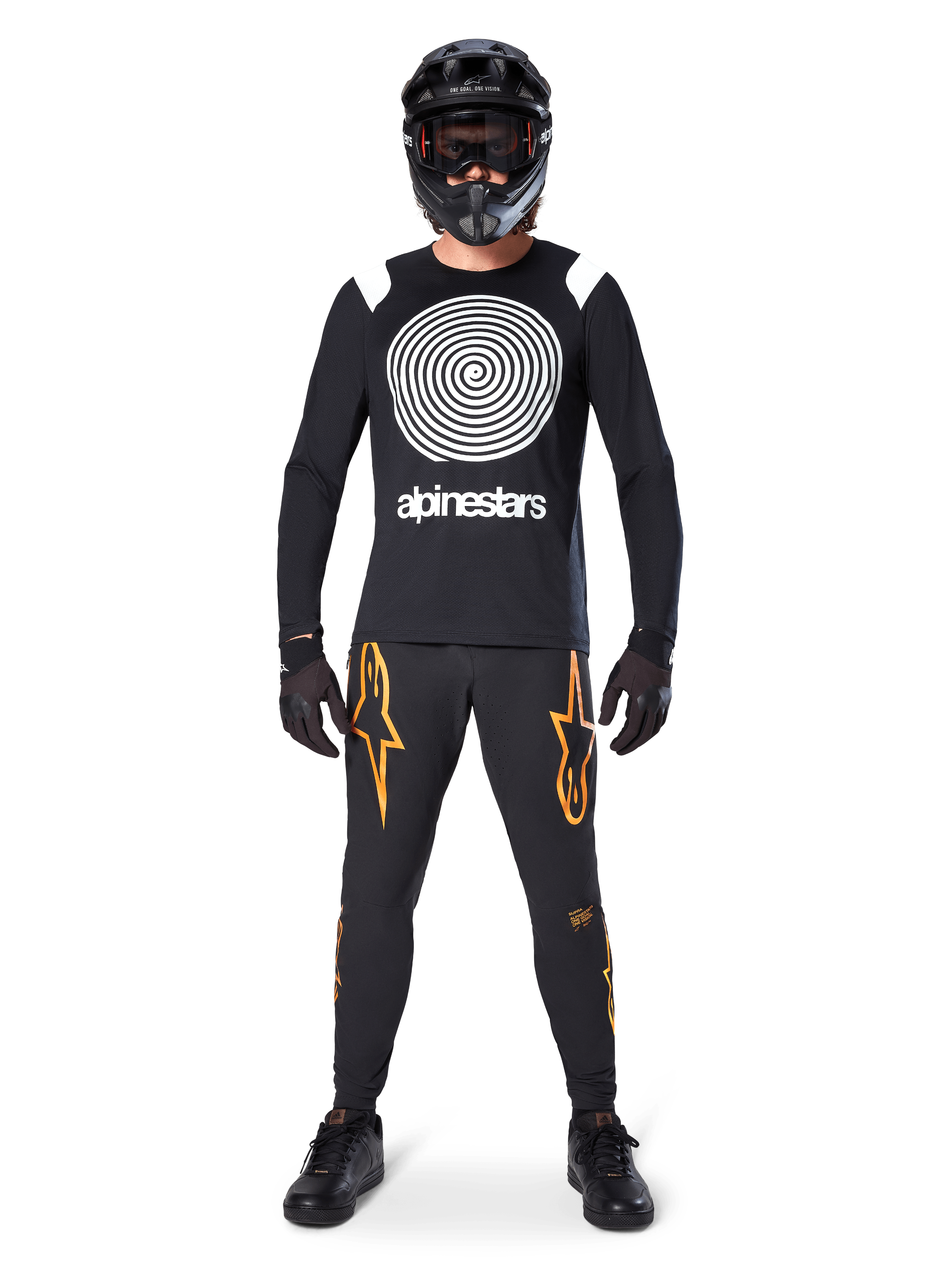 Mannelijk model in volledige mountainbike-uitrusting, Alpinestars A-Supra Oscar Jersey met lange mouwen in zwart met witte spiraalgrafiek op de borst, zwarte broek met oranje sterlogo's, zwarte full-face helm en bril, zwarte handschoenen en schoenen, gecoördineerde outfit voor MTB gravity riding