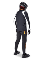 Alpinestars A-Supra Oscar Jersey met lange mouwen, zwart met witte panelen op de mouwen, gedragen door een MTB-rijder van de achterkant gezien met een zwarte full-face helm en zwarte broek met oranje logo's, fietskleding voor off-road gravity riding
