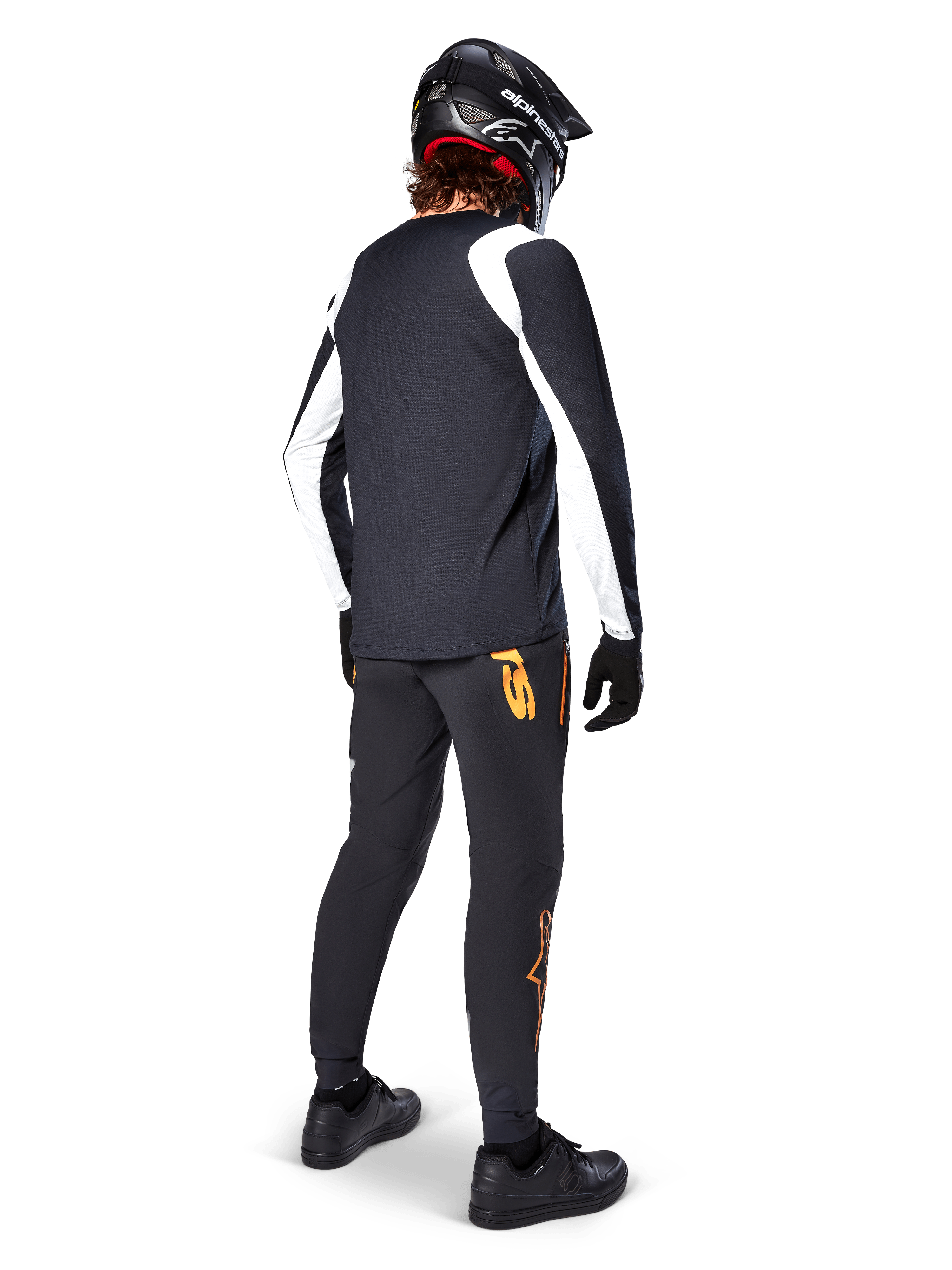 Alpinestars A-Supra Oscar Jersey met lange mouwen, zwart met witte panelen op de mouwen, gedragen door een MTB-rijder van de achterkant gezien met een zwarte full-face helm en zwarte broek met oranje logo's, fietskleding voor off-road gravity riding