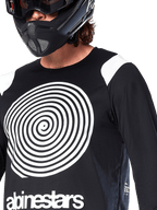 Alpinestars A-Supra Oscar Jersey, lange mouwen, zwart, met een grote witte Oscar spiraalgrafiek op de borst en merklogo, gedragen door een rijder met een zwarte full-face helm en bril, prestatiegerichte mountainbikekleding voor gravity riding