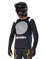 Alpinestars A-Supra Oscar Jersey met lange mouwen, MTB-fietsshirt, zwart en wit, voorzien van een opvallend wit spiraalontwerp en Alpinestars-logo op de borst, gedragen met een zwarte full-face helm en bril, prestatiegerichte mountainbikekleding