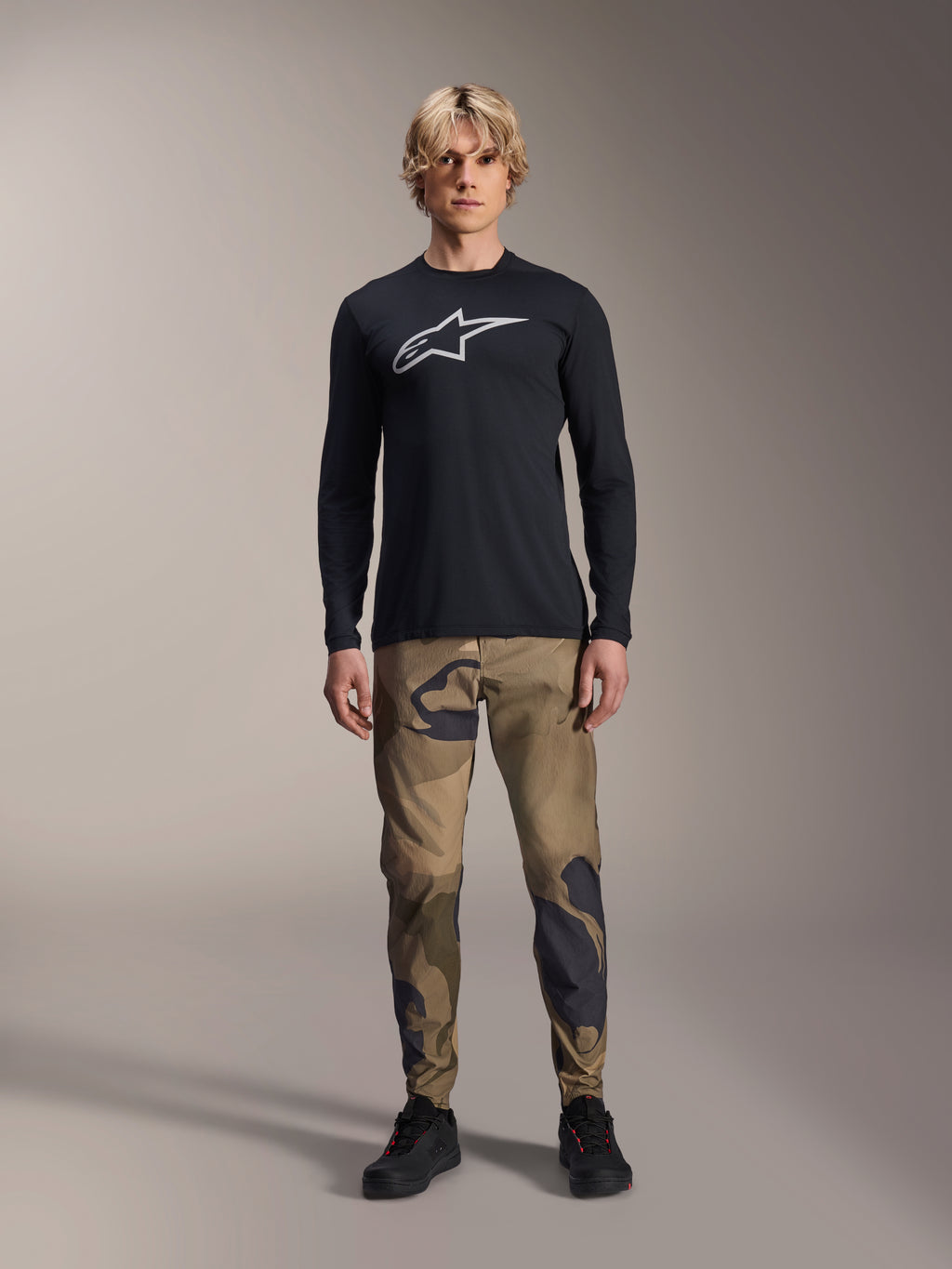 Alpinestars A-Dura Camo Pant, MTB-fietsbroek, bruin en groen camouflagepatroon, gedragen met een zwart shirt met lange mouwen en zwarte schoenen, sportieve mountainbike-stijl met duurzame stretchstof