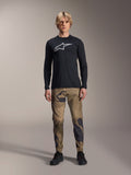 Alpinestars A-Dura Camo Pant, MTB-fietsbroek, bruin en groen camouflagepatroon, gedragen met een zwart shirt met lange mouwen en zwarte schoenen, sportieve mountainbike-stijl met duurzame stretchstof
