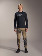 Alpinestars A-Dura Camo Pant, MTB-fietsbroek, bruin en groen camouflagepatroon, gedragen met een zwart shirt met lange mouwen en zwarte schoenen, sportieve mountainbike-stijl met duurzame stretchstof