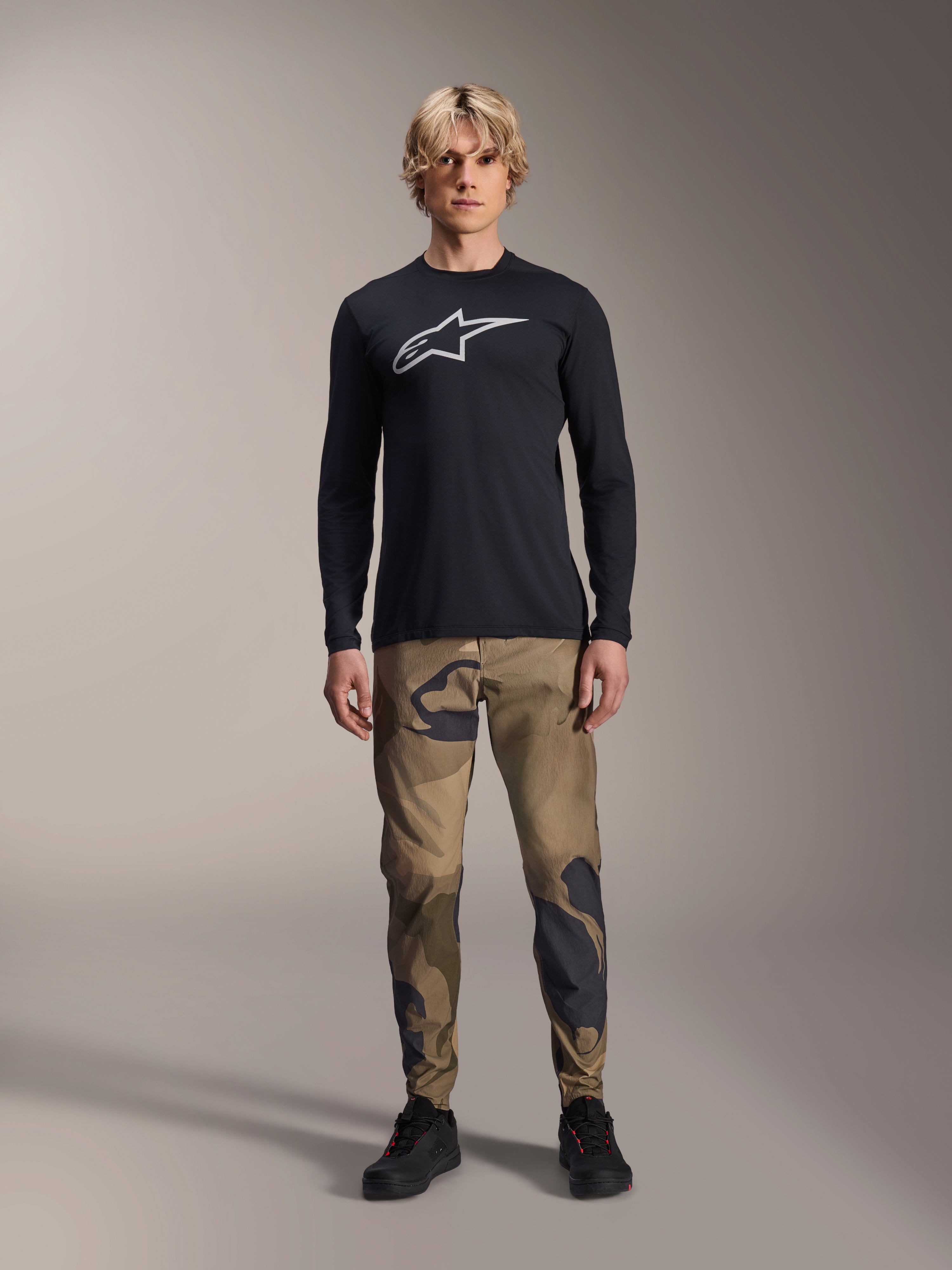 Alpinestars A-Dura Camo Pant, MTB-fietsbroek, bruin en groen camouflagepatroon, gedragen met een zwart shirt met lange mouwen en zwarte schoenen, sportieve mountainbike-stijl met duurzame stretchstof